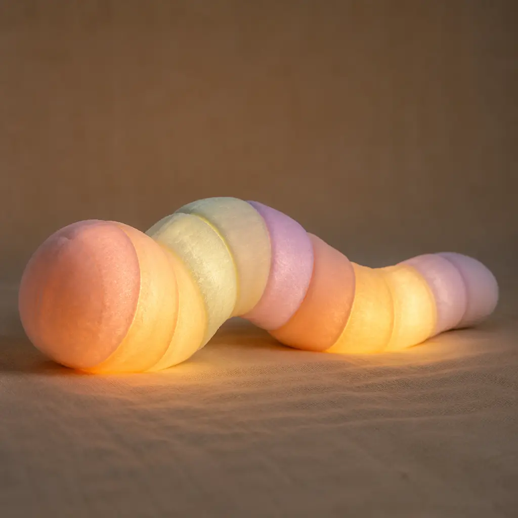A softly glowing plush worm toy resting on a bed in low light